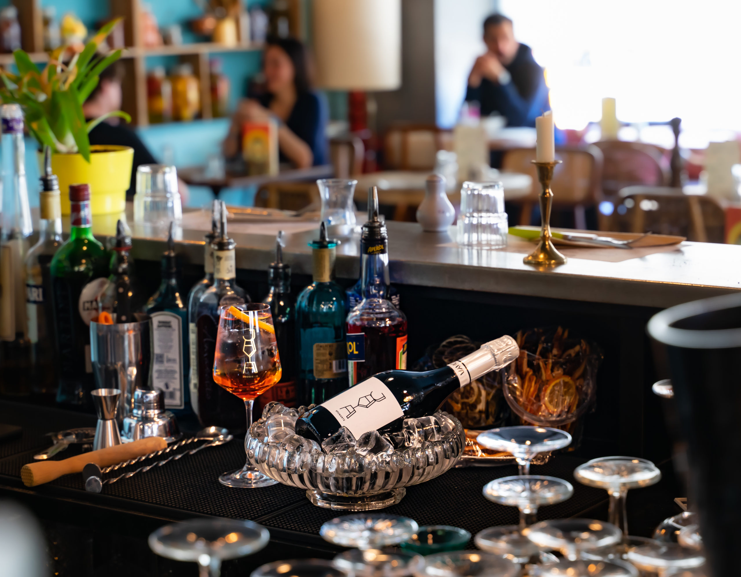 Photographie montrant une bouteille de JAYA Spumante et un Apérol Spritz posé sur un bar avec, en arrière-plan, des gens assis à des tables.